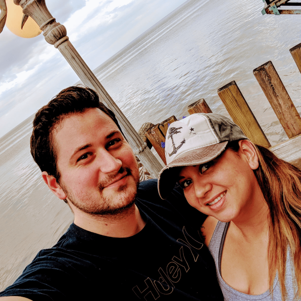 Amanda and Nathan in Cedar Keys, FL with the ocean behind them. Amanda is wearing a gray and white baseball cap and a gray shirt. Nathan is wearing a dark blue shirt.